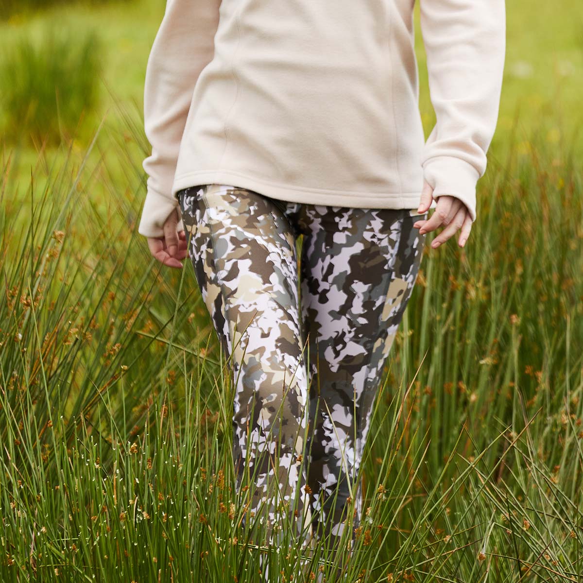 Person wearing camouflage pants and a light-colored jacket standing in tall grass.