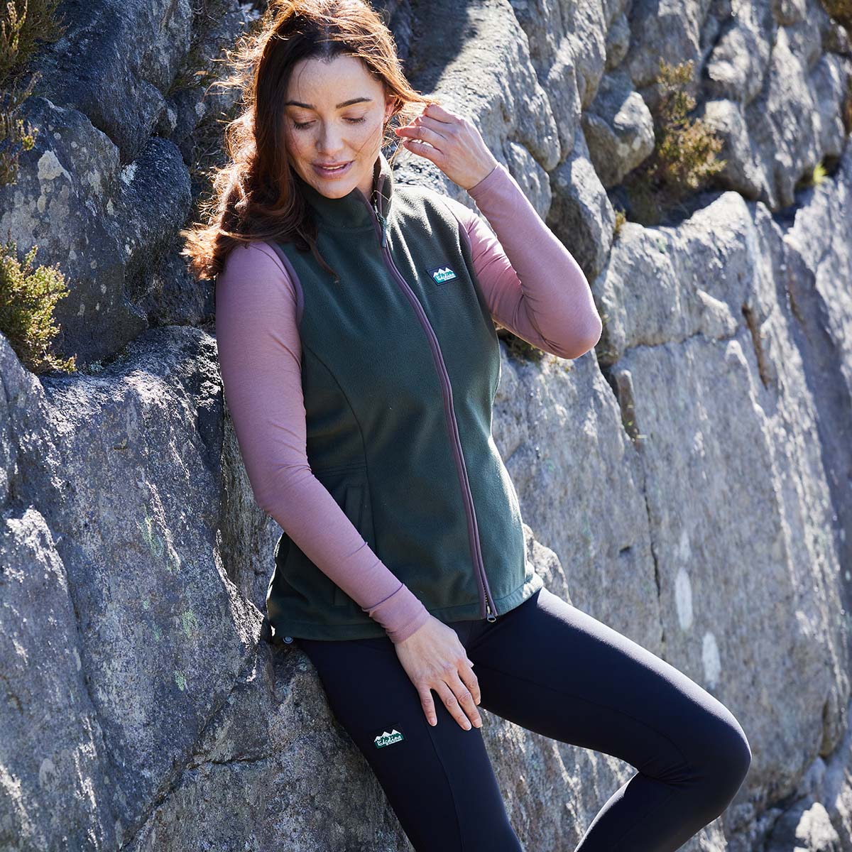 Woman sitting on a rocky surface wearing a green vest and black leggings with a visible Ridgeline Clothing logo.