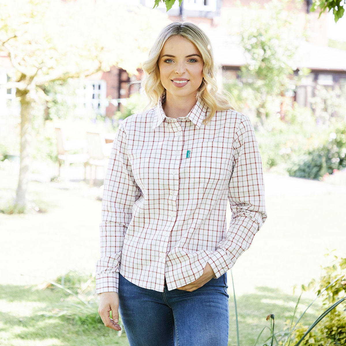 Woman wearing a checkered shirt and jeans standing outdoors with greenery in the background