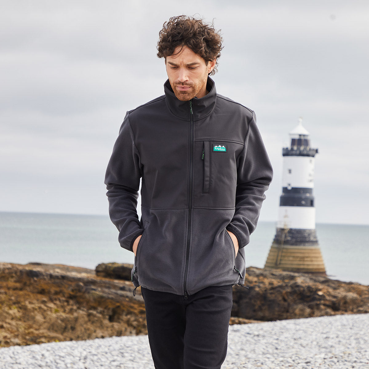 Man wearing a grey & black windproof fleece jacket with a Ridgeline Clothing logo on a rocky beach with a lighthouse in the background