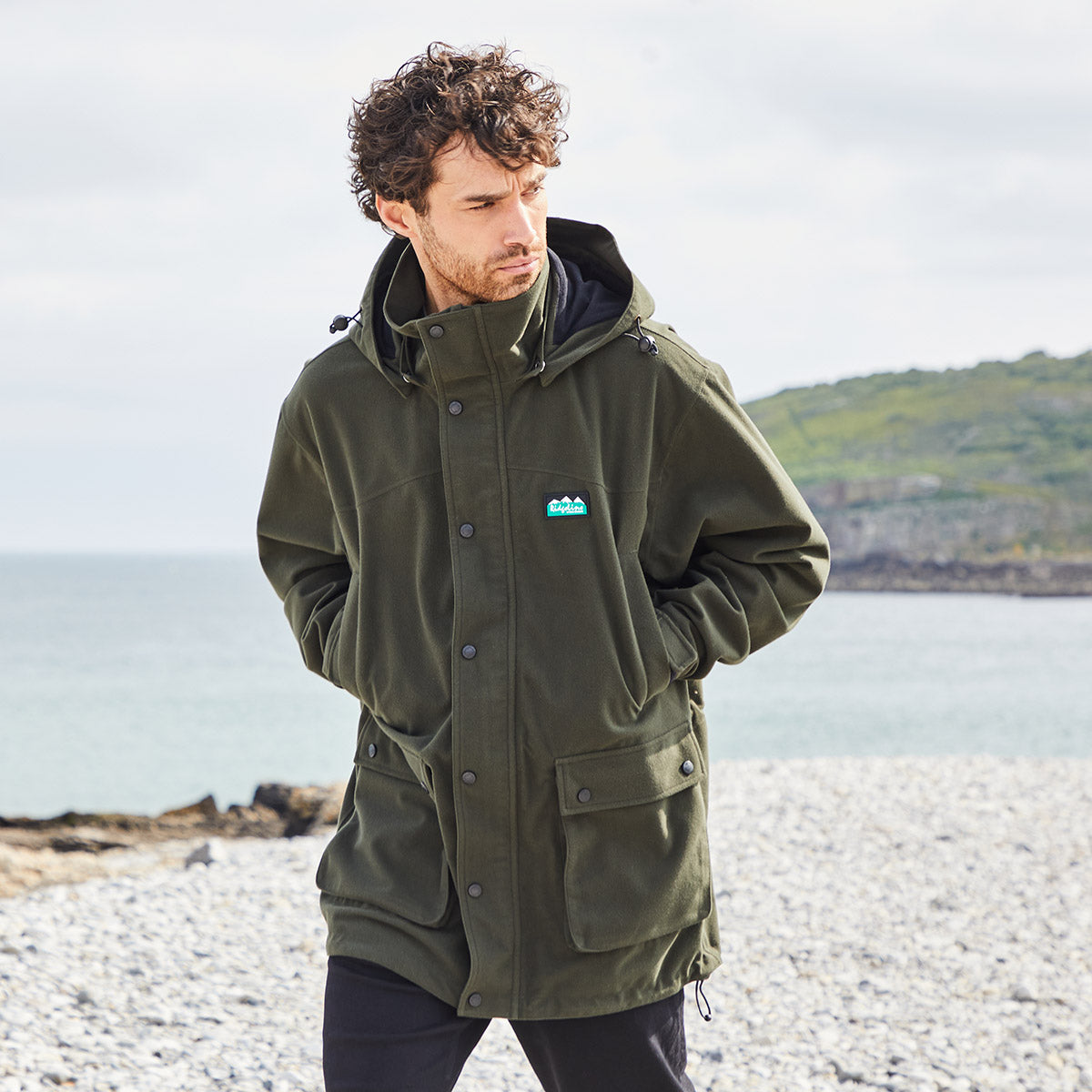 Man wearing a dark green waterproof jacket with a Ridgeline Clothing brand logo on a beach