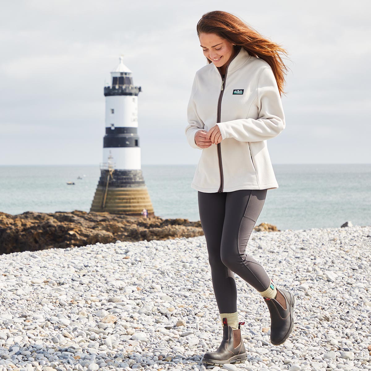 Woman walking on a pebbly beach wearing black leggings & Cream fleece jacket with a lighthouse in the background