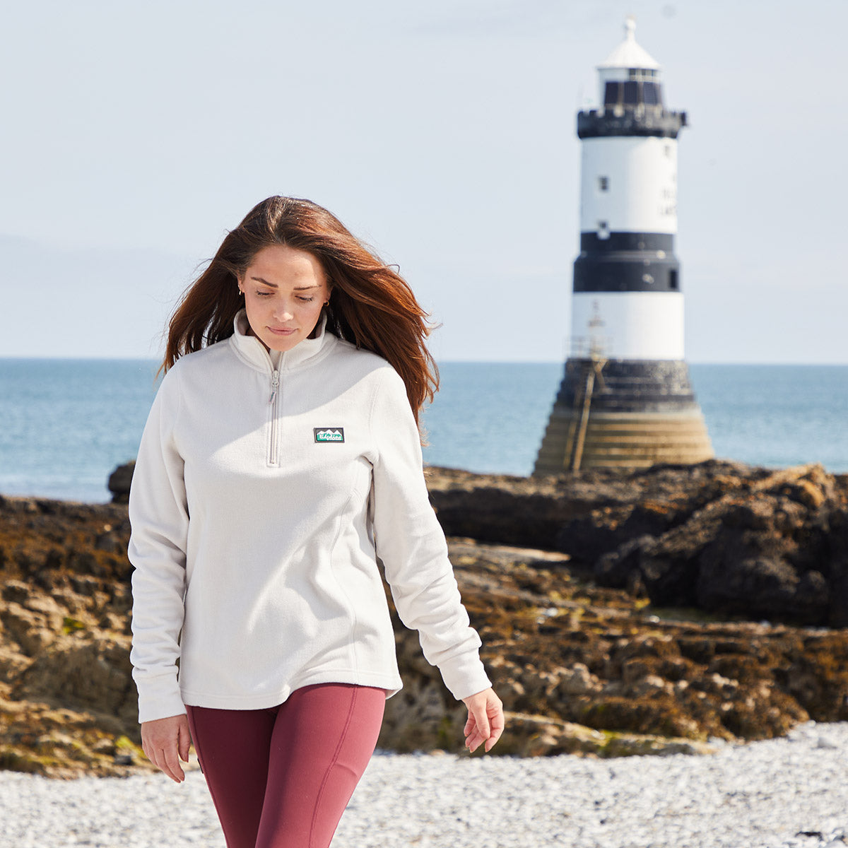 Woman walking on a pebbly beach with a lighthouse in the background. Wearing a cream fleece with Ridgeline Clothing logo on Chest