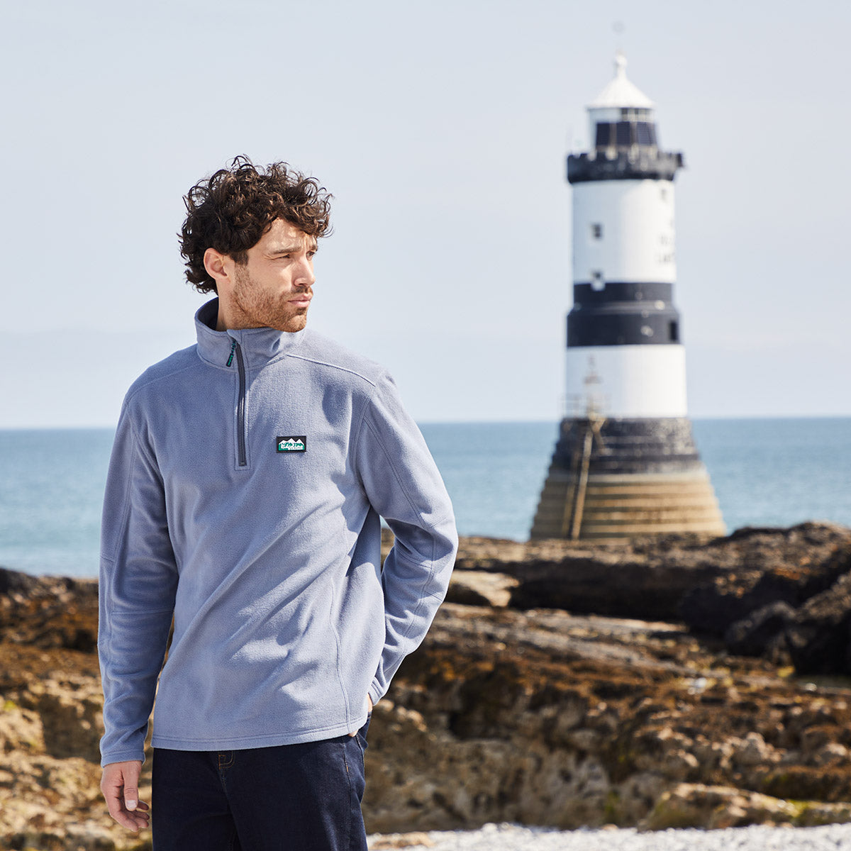 Man wearing a light blue pullover with a Ridgeline Clothing logo, standing on rocks with a lighthouse in the background.