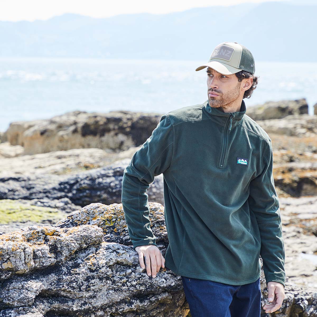 Man wearing a green windproof fleece and cap standing on rocky terrain with mountains in the background
