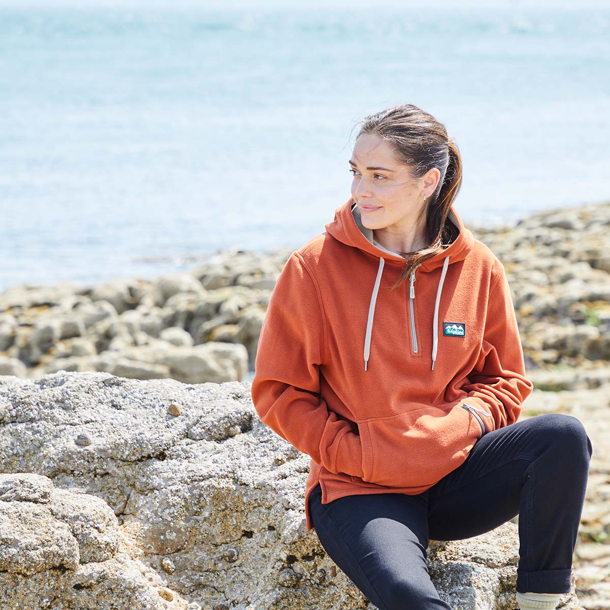 Person wearing an orange fleece hoodie with a Ridgeline Clothing logo, sitting on rocks by the sea.