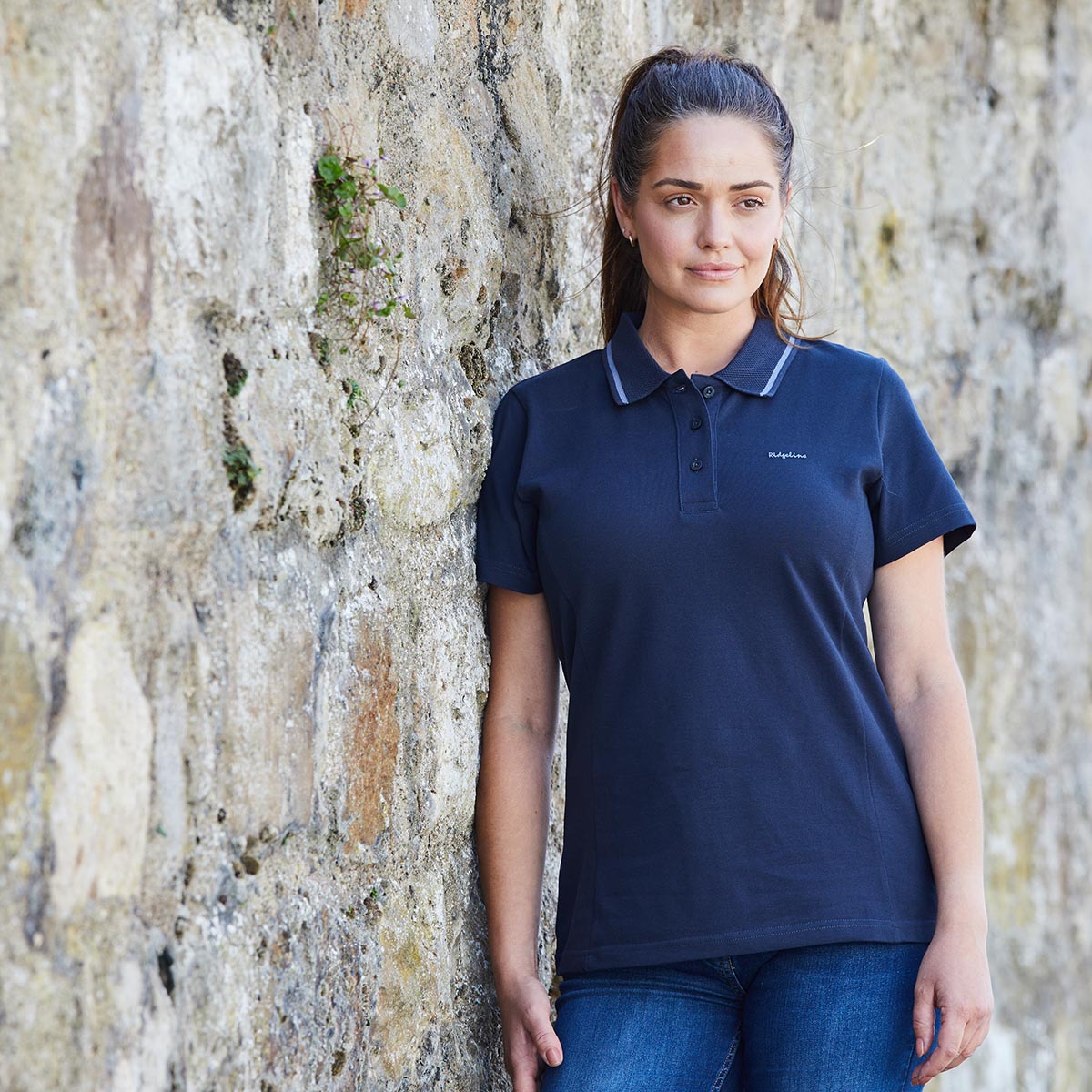 Woman wearing a navy polo shirt standing against a stone wall.