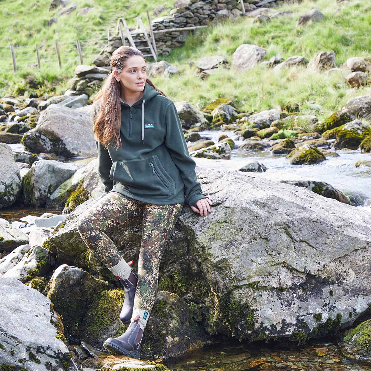 Woman in Green fleece hoodie with kangaroo pockets & Ridgeline clothing logo on chest. Sitting on a rock by a stream