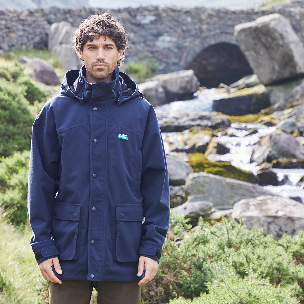 Man wearing a blue waterproof jacket with a Ridgeline Clothing logo standing in a natural setting with rocks and water.