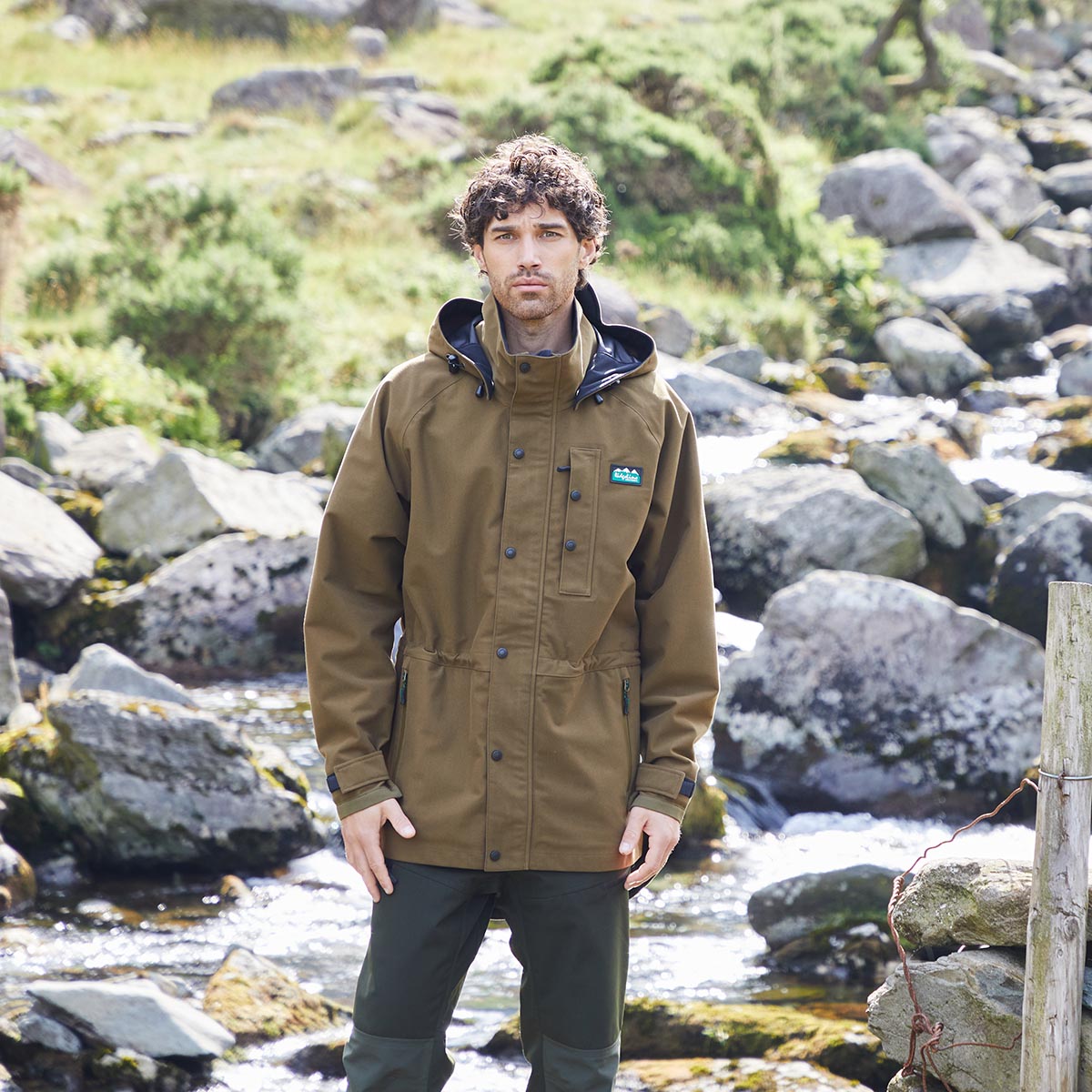 Man wearing a brown waterproof jacket standing in a natural setting with rocks and water.