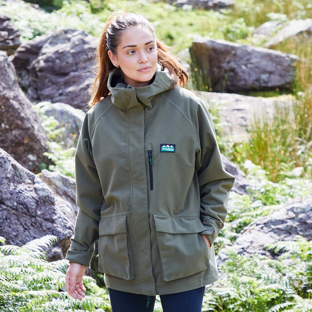 Woman wearing a green waterproof jacket with a Ridgeline Clothing logo in a natural setting