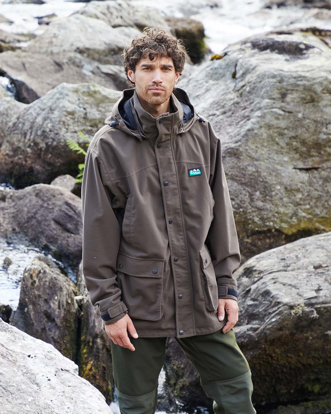 Man wearing a brown jacket with a logo standing in a rocky outdoor setting