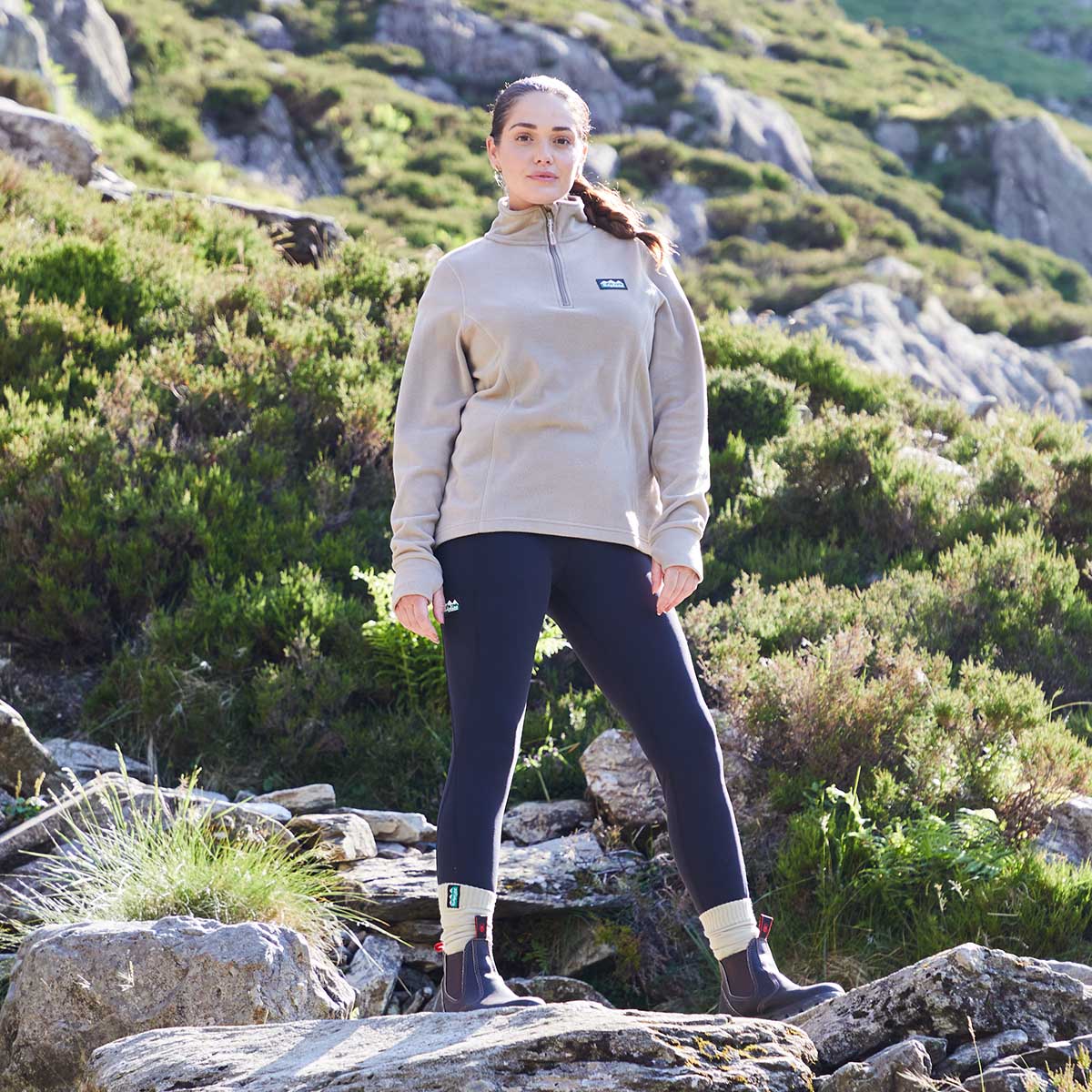 Woman standing on a rocky hillside wearing a beige pullover and dark blue leggings.