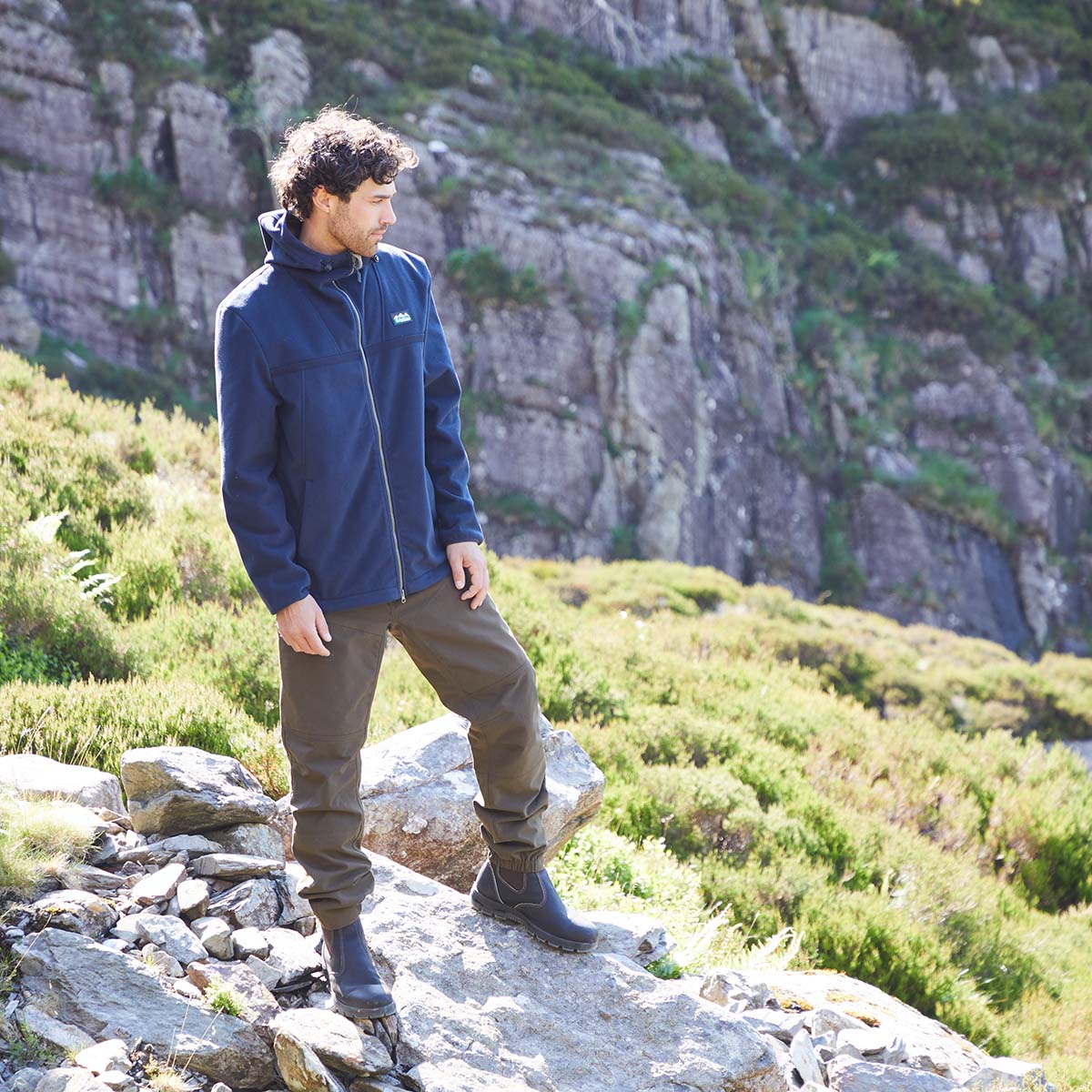 Man in outdoor clothing standing on a rocky mountain path with a mountainous background