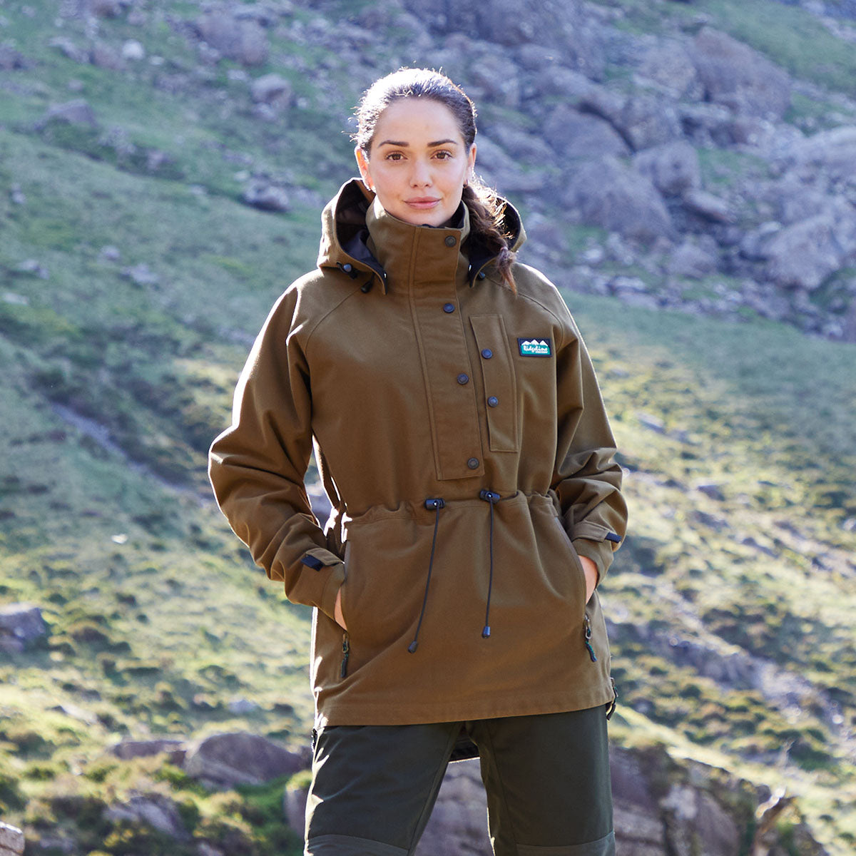 Woman wearing a brown waterproof jacket with a Ridgeline Clothing logo in a mountainous landscape