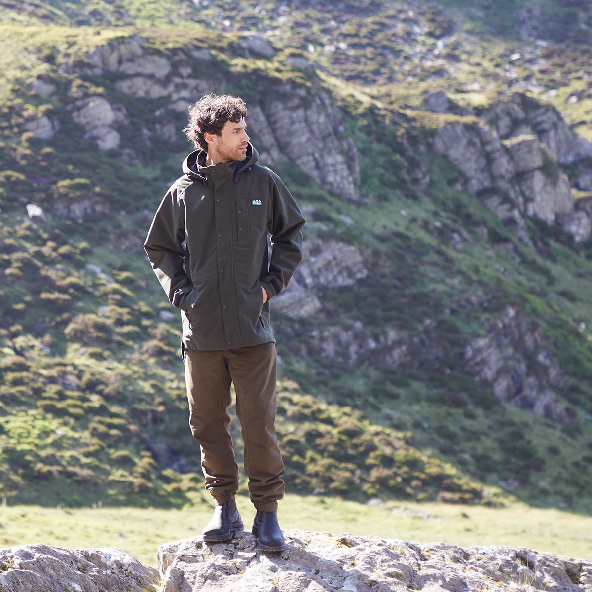 Person wearing a green jacket and brown pants standing on a rocky outcrop with a mountainous landscape in the background.