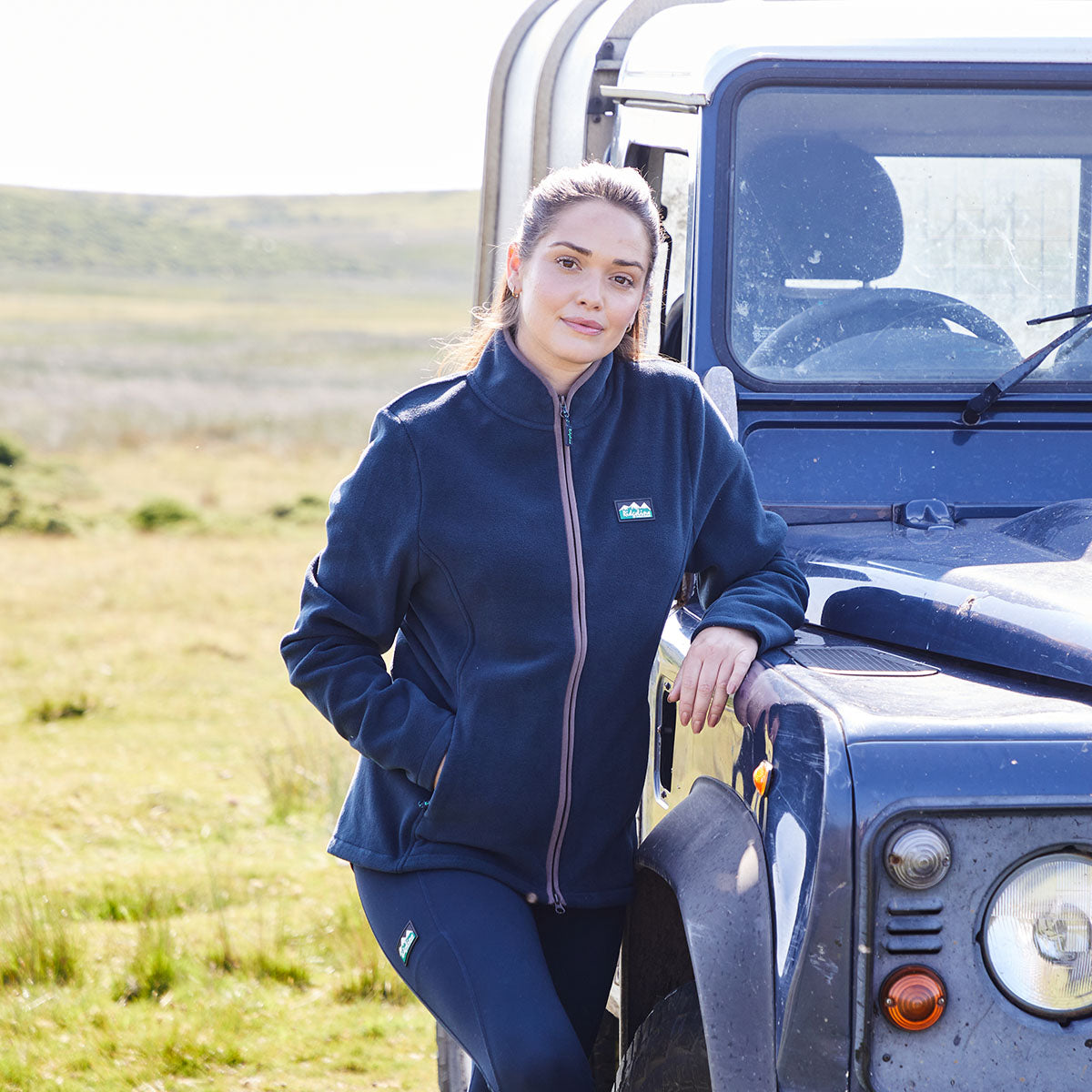 Woman in a navy blue windproof fleece jacket standing next to a Land Rover in a field