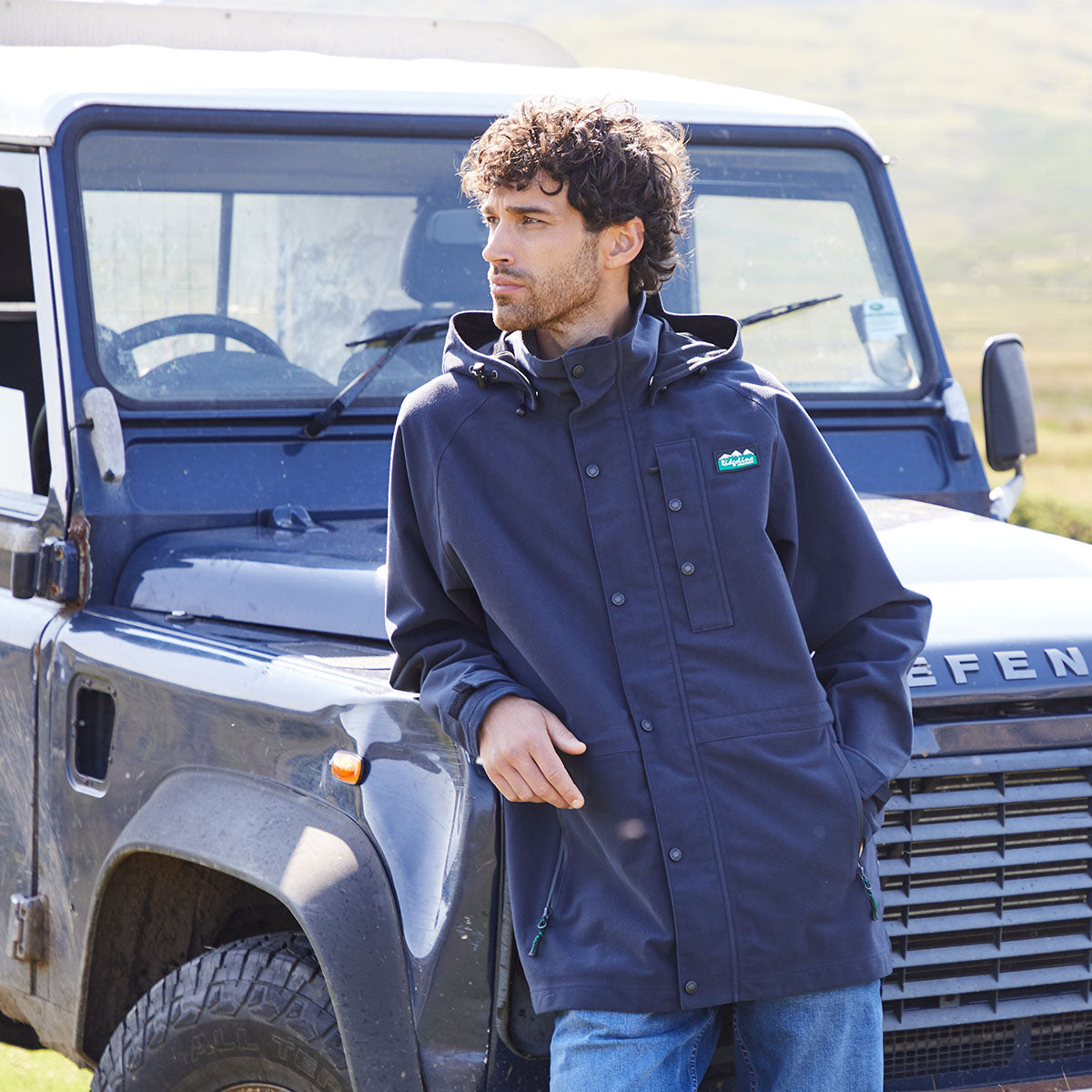 Man wearing a blue waterproof jacket standing next to a Land Rover Defender in a field.
