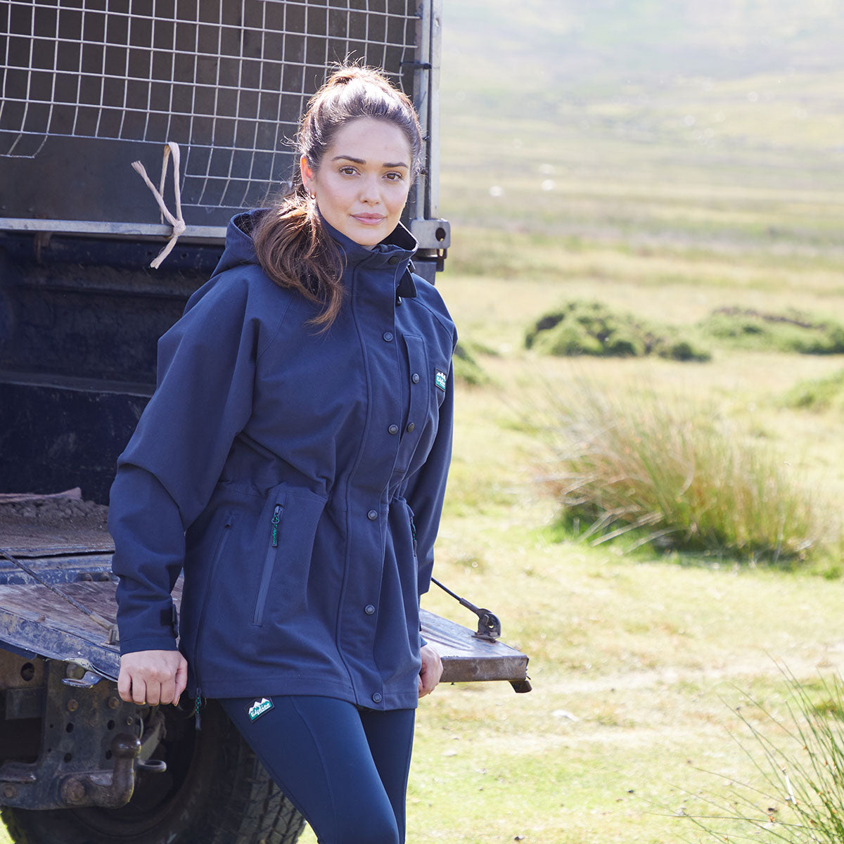Woman wearing a blue waterproof jacket standing next to a truck in a grassy field