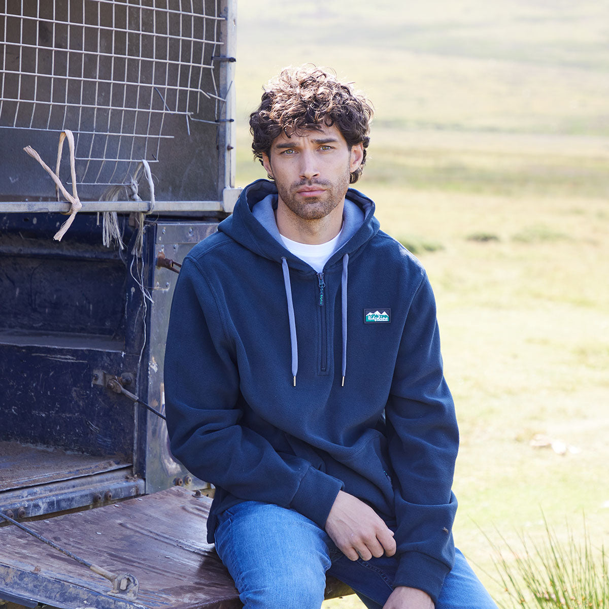 man wearing navy hoodie with a Ridgeline clothing logo on a white background. Showing Kangaroo pockets with a zip. sitting outdoors near a vehicle.