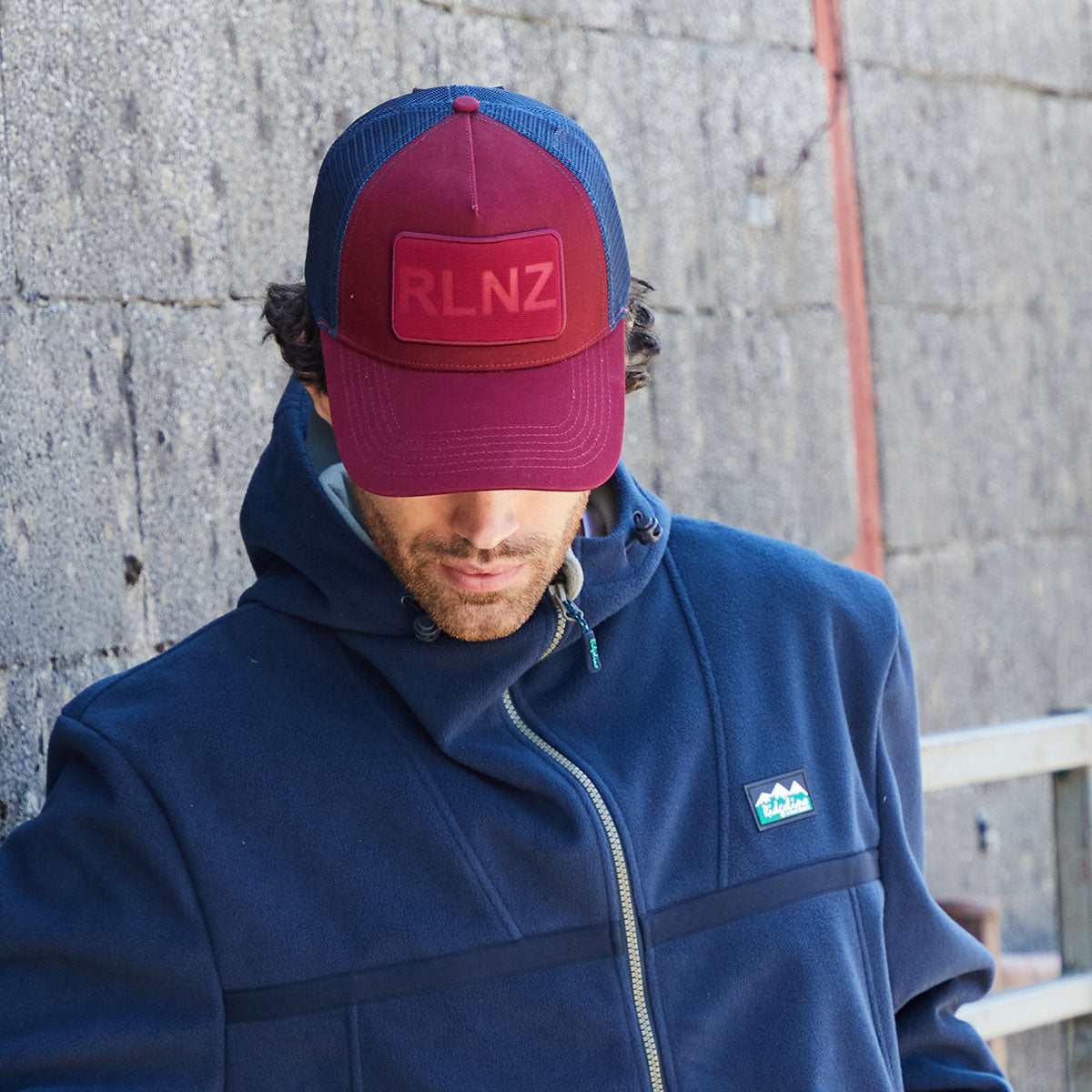 Person wearing a blue jacket with a visible brand logo and a maroon and blue cap with 'RLNZ' on it, standing against a grey wall.