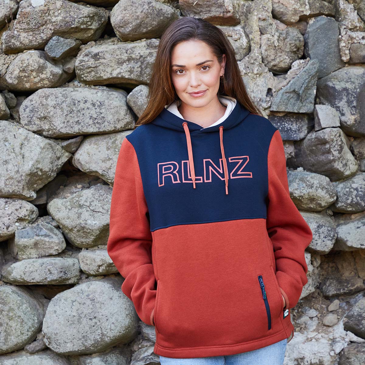 Person wearing a red and navy hoodie with 'RLNZ' logo against a stone wall.