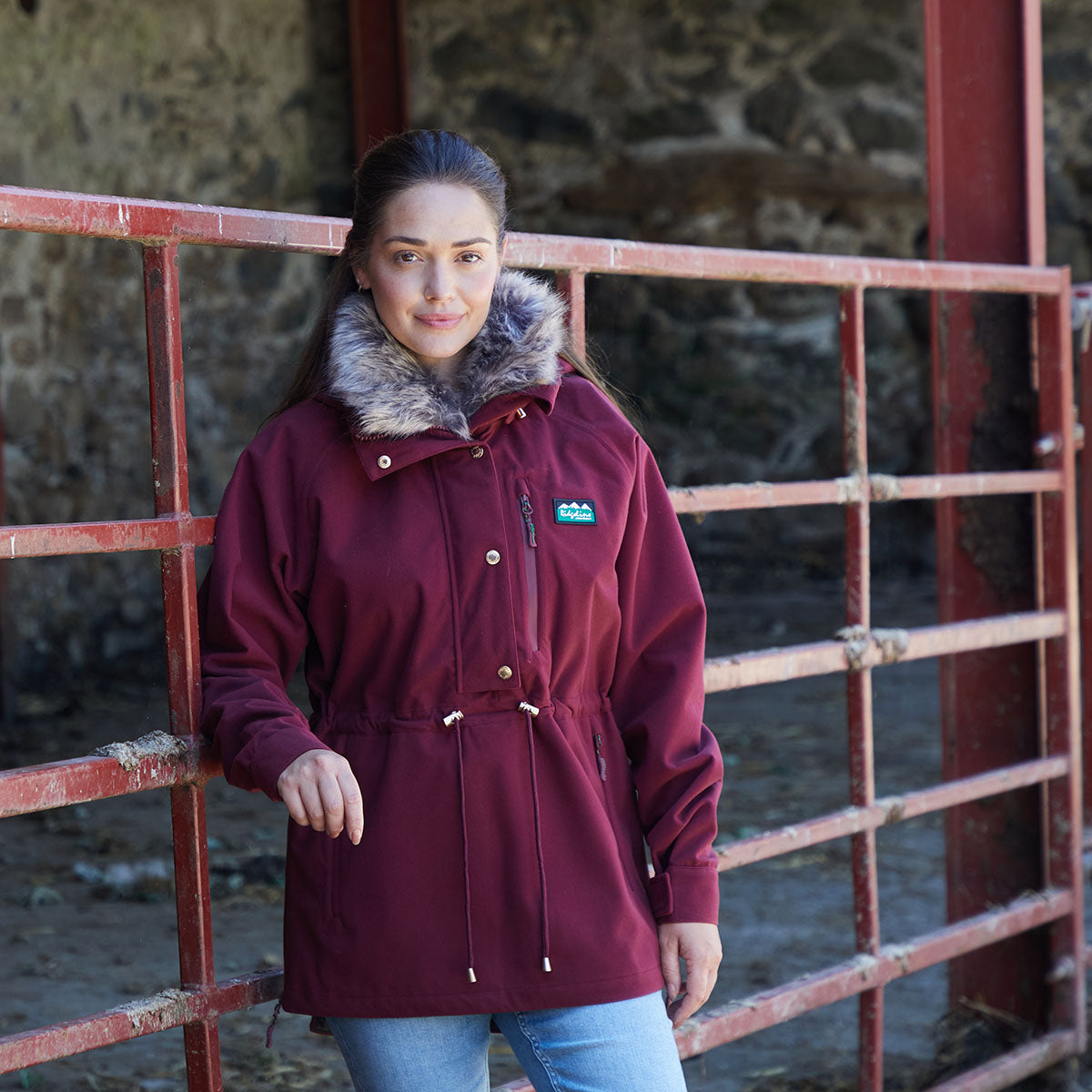 Woman wearing a maroon coat with fur-lined hood in a barn setting leaning on a metal fence