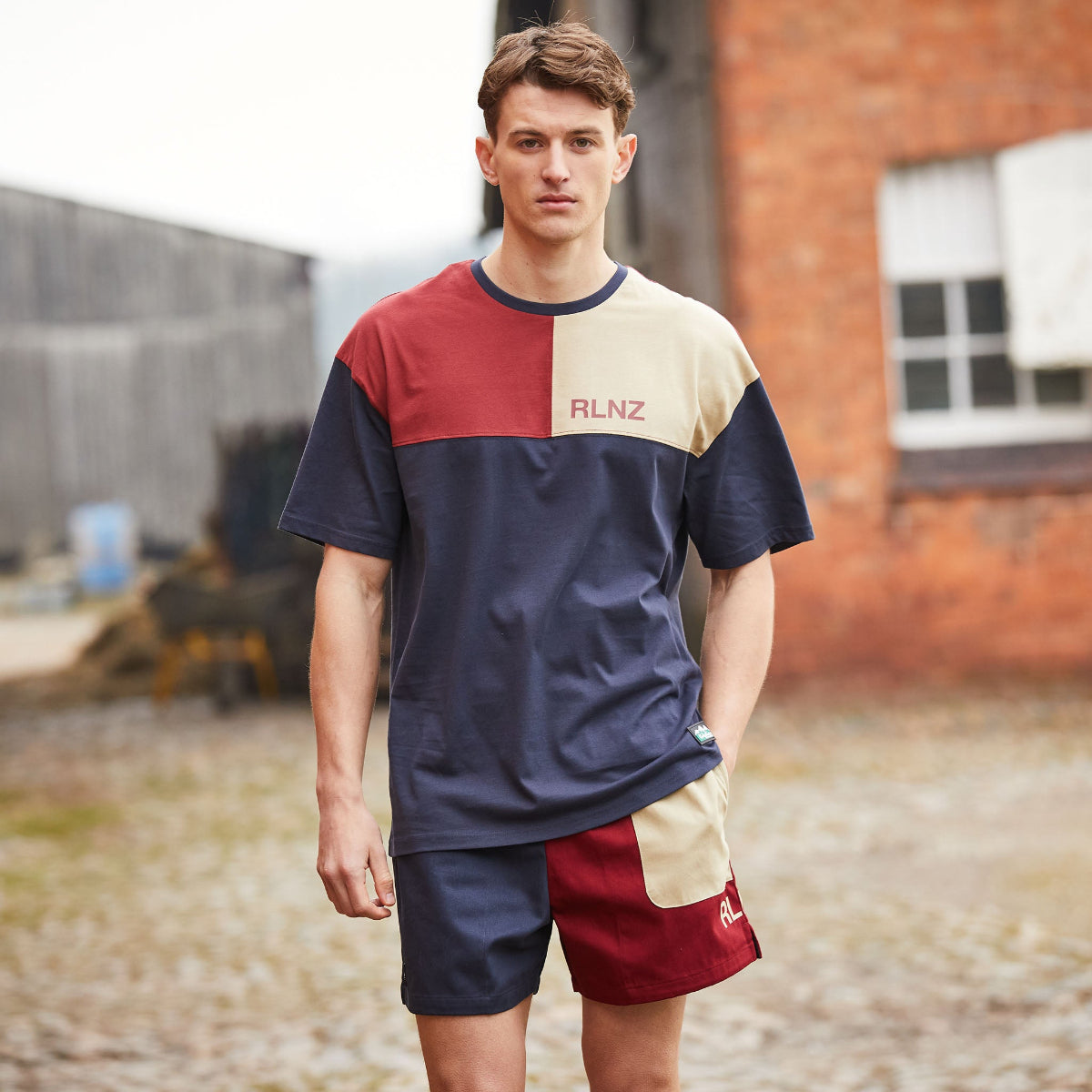 Man wearing a color-blocked shirt and shorts with 'RLNZ' branding outdoors. Standing infront of a house