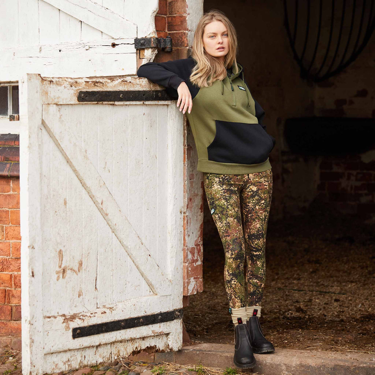 Woman wearing a pair of camouflage patterned leggings with a high waist and side pockets. Leaning against horse stable