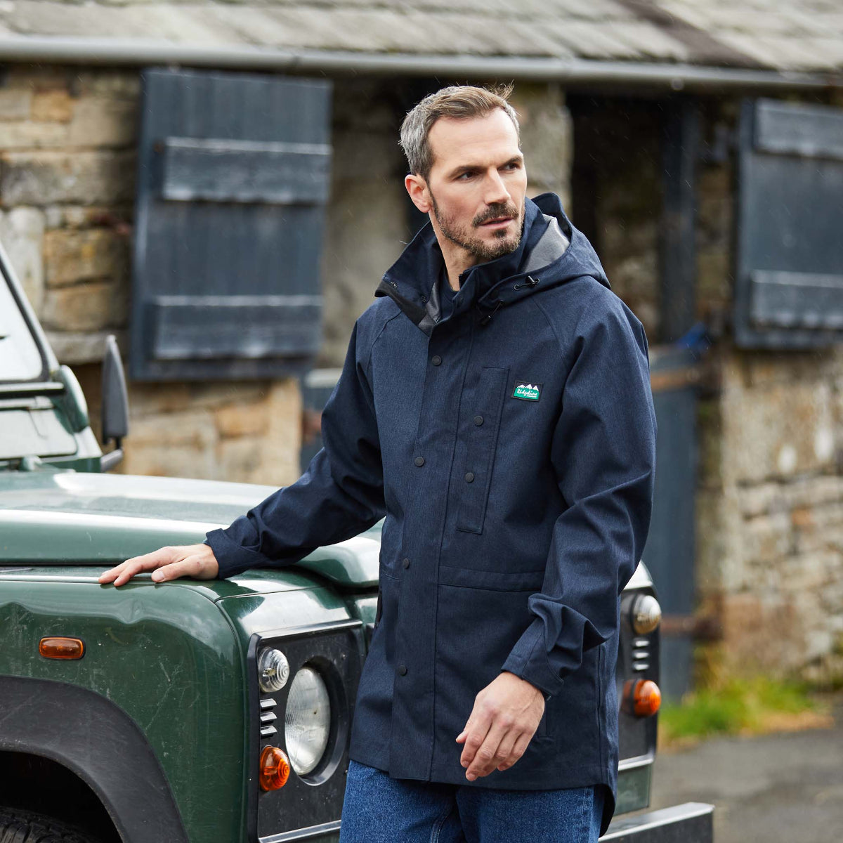 Man wearing a Navy Monsoon light waterproof jacket Windproof with a hood, featuring a full-length front zip, two zipped hand pockets, and a zipped chest pocket. standing & leaning next to a jeep