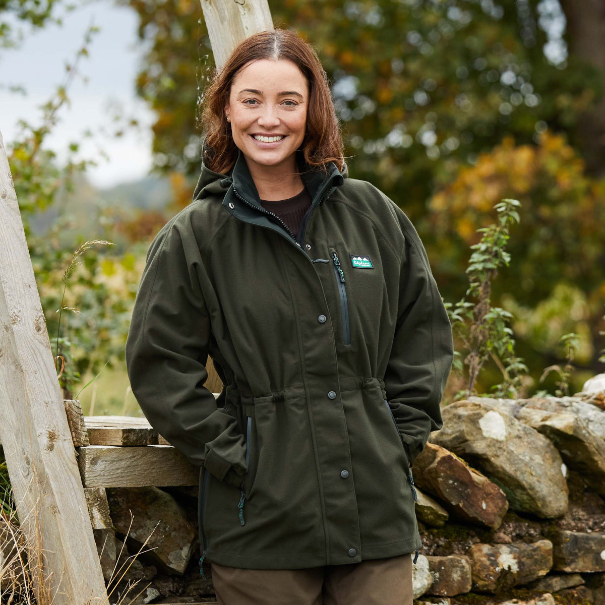 woman wearing a A green full-zip waterproof jacket with a hood, featuring a chest pocket and side hip zips, designed for outdoor use. leaning on a stone wall