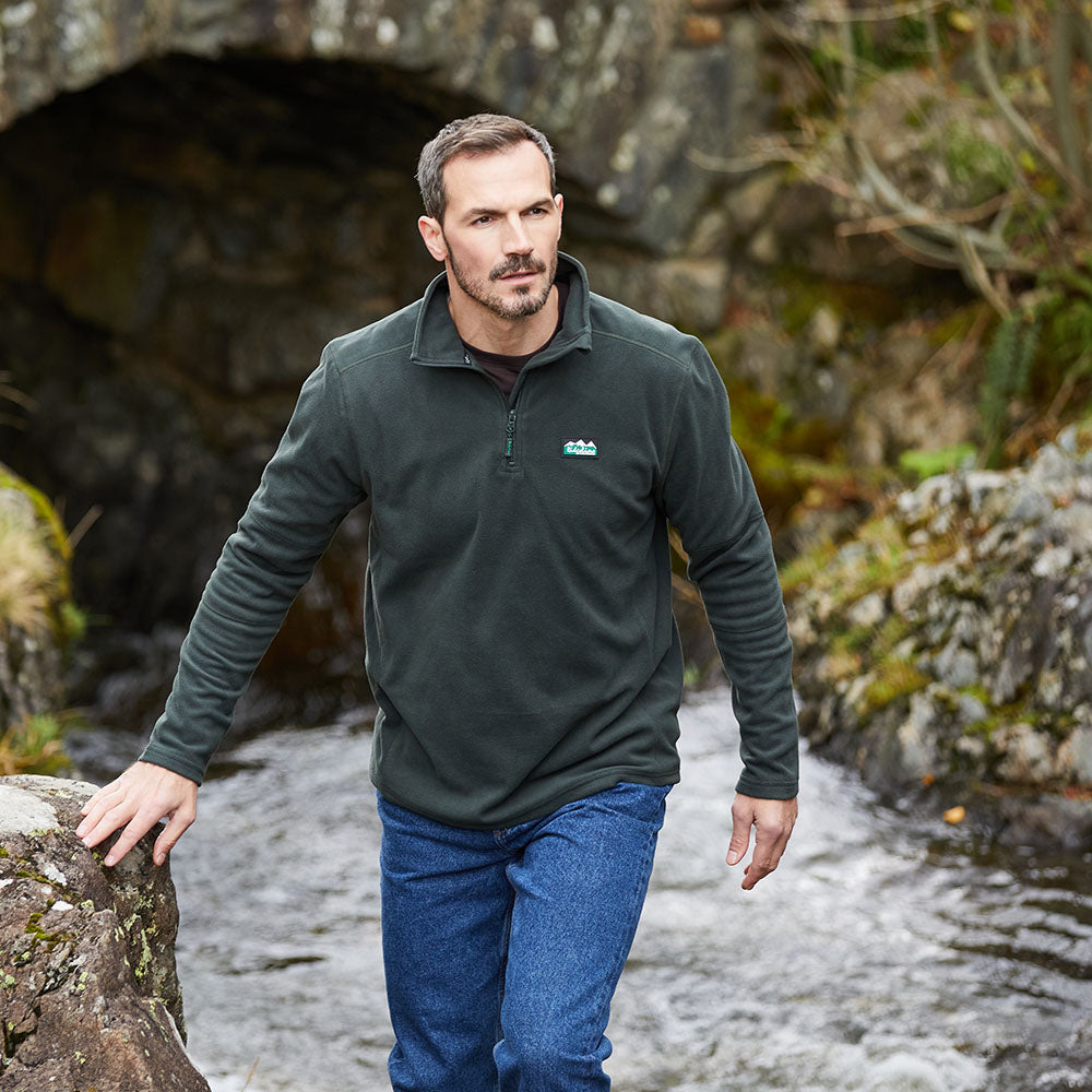 A man wearing green windproof fleece pullover with a quarter zipper and a Ridgeline Clothing logo on the chest with river behind him