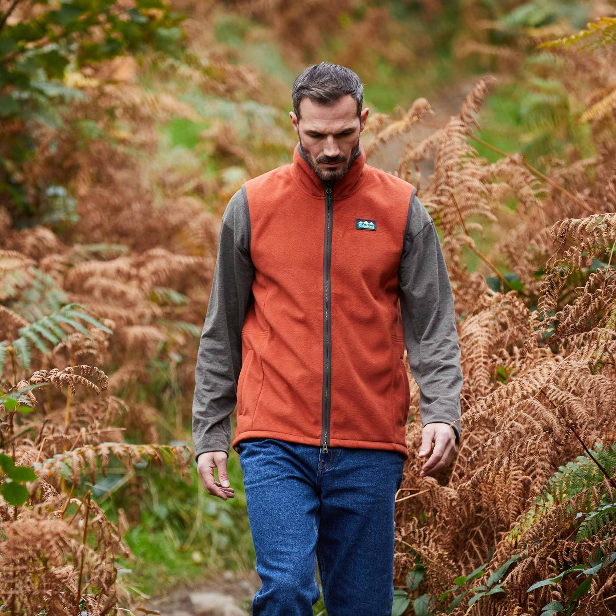 Man walking through a forest wearing an orange vest with a Ridgeline Clothing brand logo. Walking through high plants