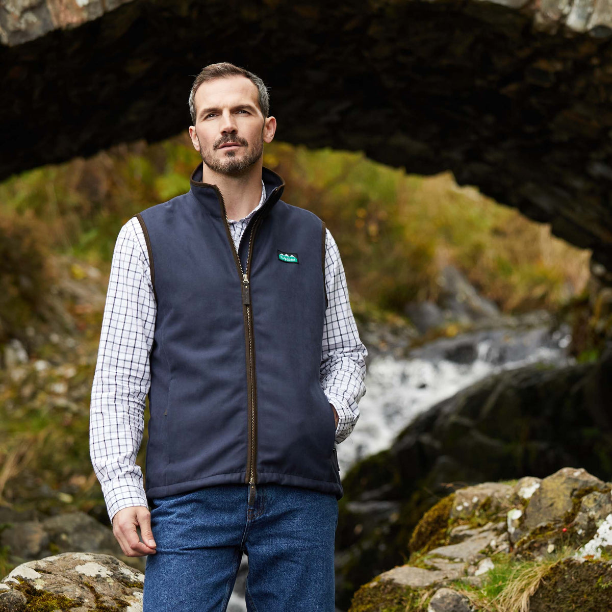 Man wearing a navy gilet with a logo standing in front of a natural landscape with rocks and water.