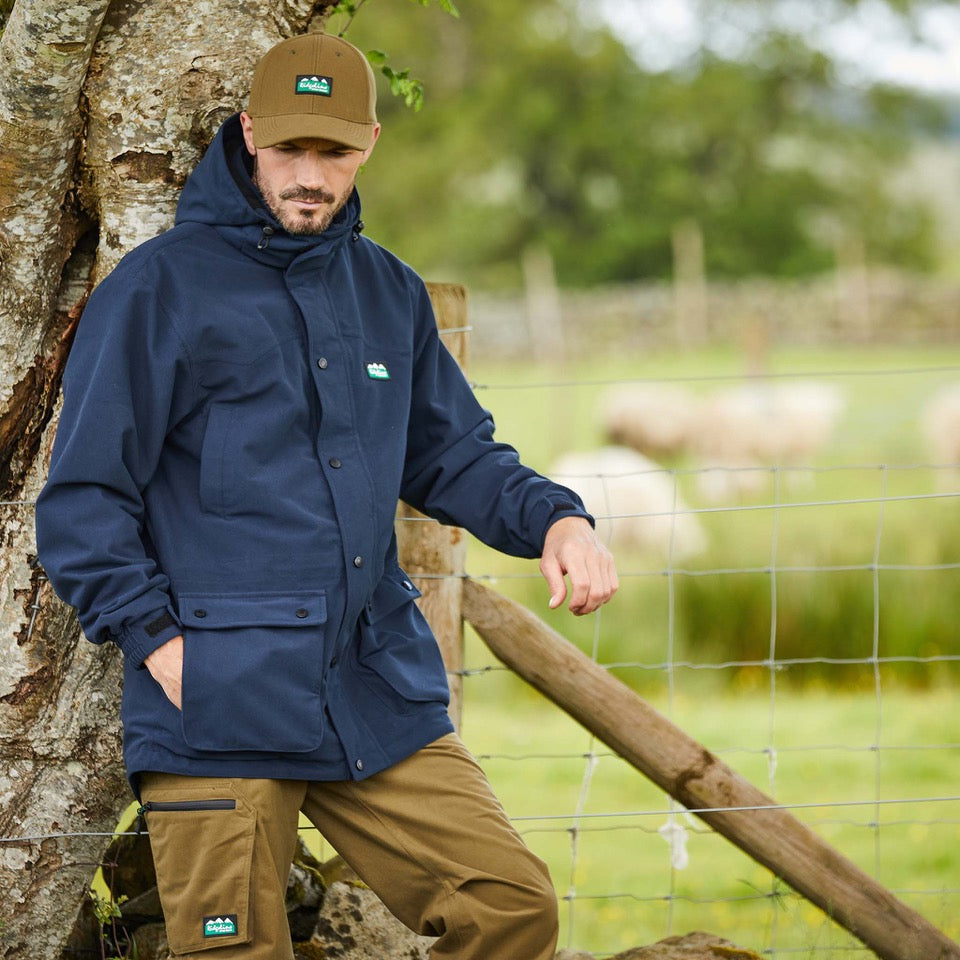 Man wearing a navy-coloured Ridgeline Torrent III Jacket with a detachable hood, zip front, and multiple pockets. leaning on a metal fence with a tree behind him