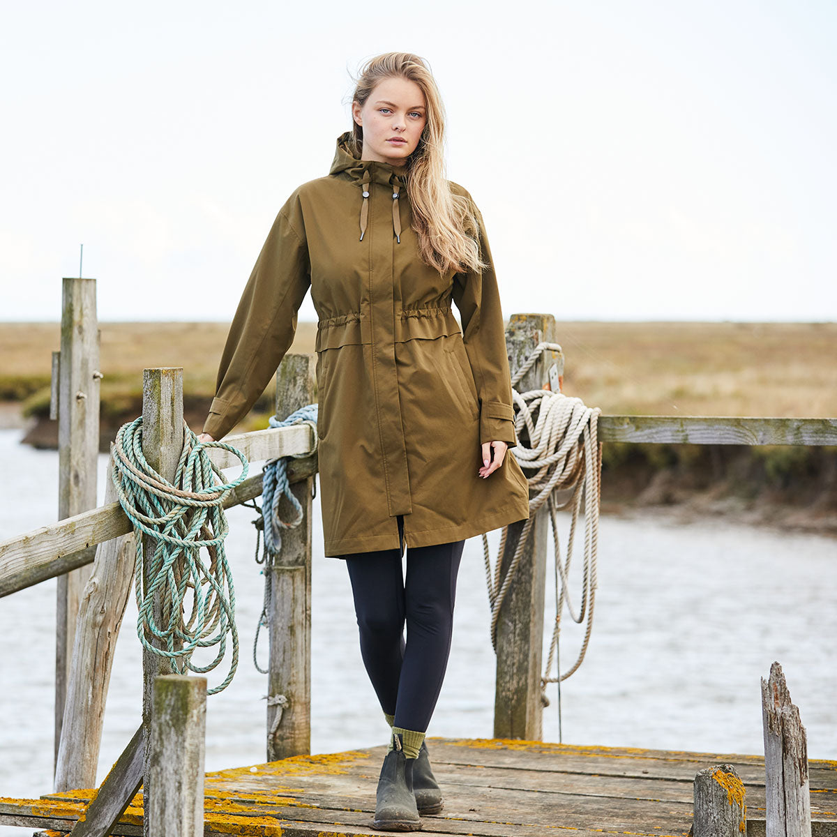 Woman wearing a brown raincoat standing on wooden deck by the sea