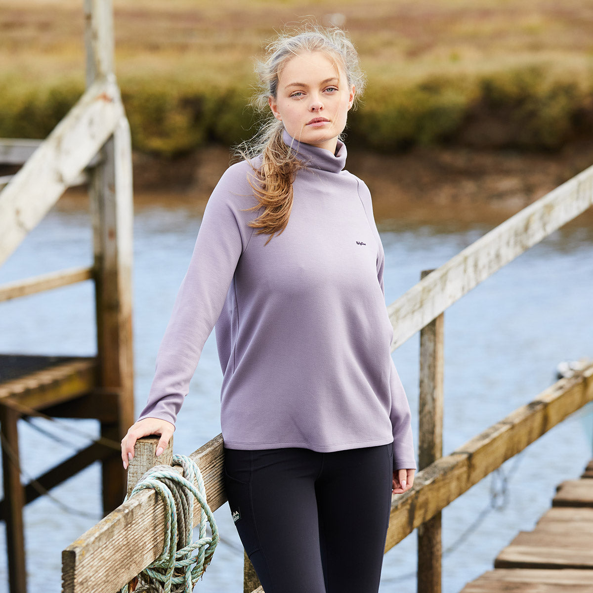 Woman wearing a light purple turtleneck sweater and black leggings standing on a wooden bridge over water.