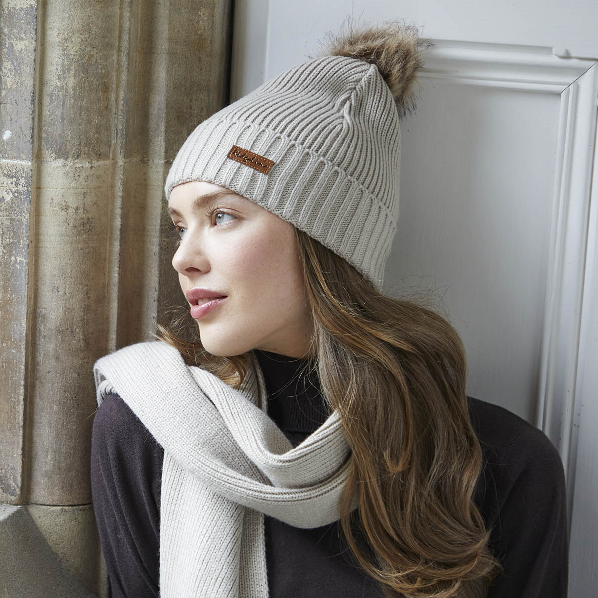 Model wearing cream ribbed knit beanie and scarf set with faux fur pom pom, by the window.