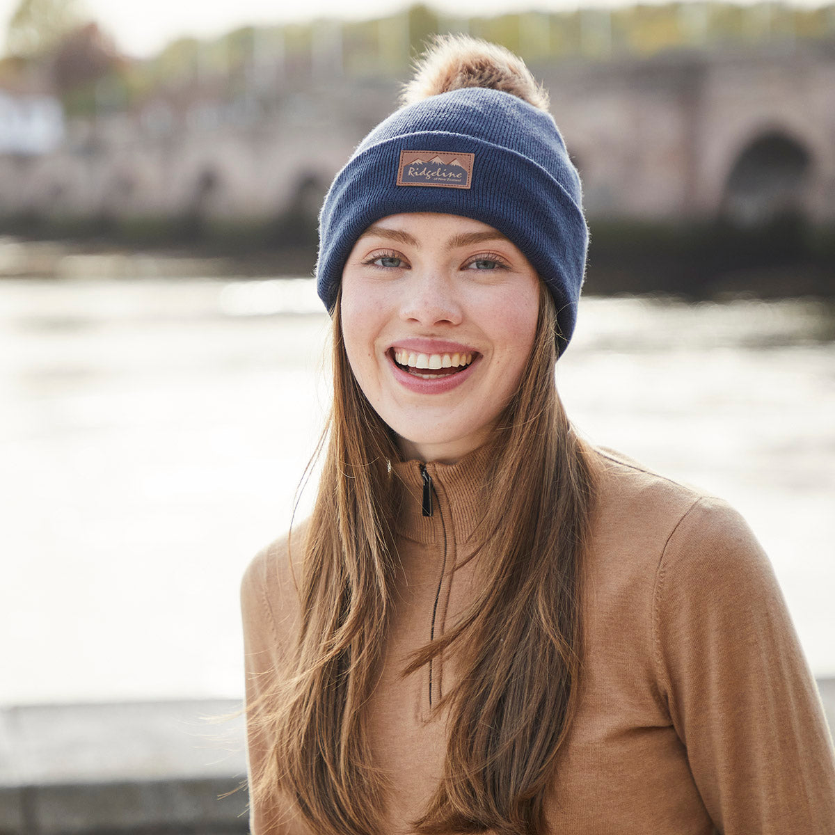 Lifestyle image of a person wearing a navy blue ribbed knit beanie with a folded cuff and a large faux-fur pom-pom on the crown. A brown faux-leather Ridgeline of New Zealand logo patch is displayed on the front cuff. The person is dressed in a light brown quarter-zip top and photographed outdoors near a body of water, with a softly blurred stone bridge visible in the background. The image highlights the beanie's soft knit texture, comfortable fit, and stylish cold-weather design.
