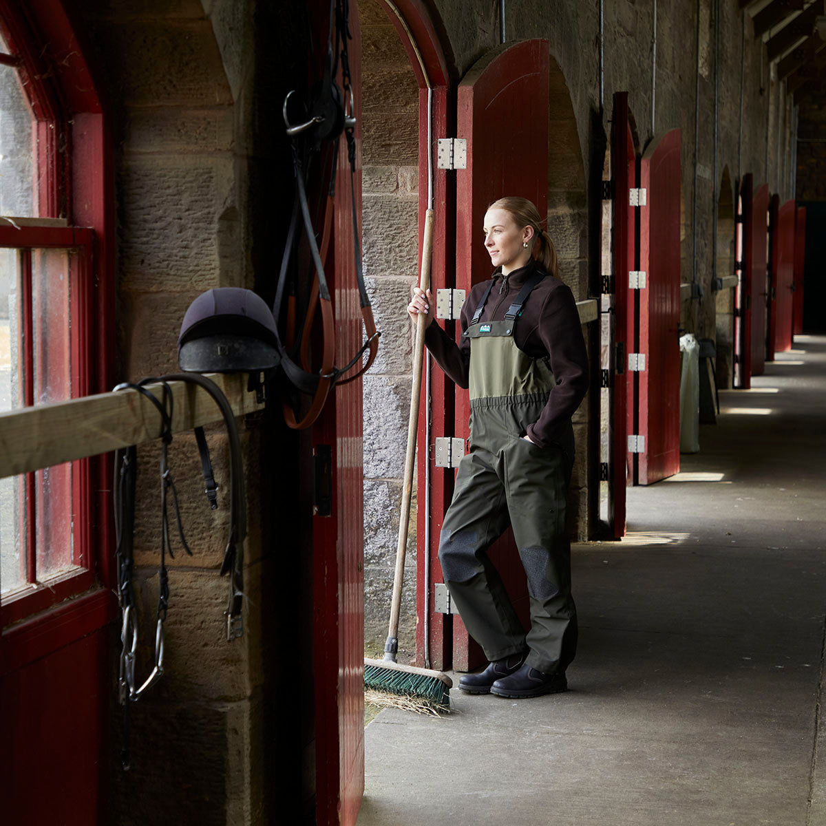 Lifestyle image of a person wearing olive-green waterproof bib and brace trousers while standing in a stable corridor. The bib and brace features adjustable shoulder straps, reinforced knee panels, and integrated waterproof boots designed for outdoor and equestrian environments. The person is holding a yard broom beside an open stable door, with multiple red stable doors lining the stone-built passageway in the background.