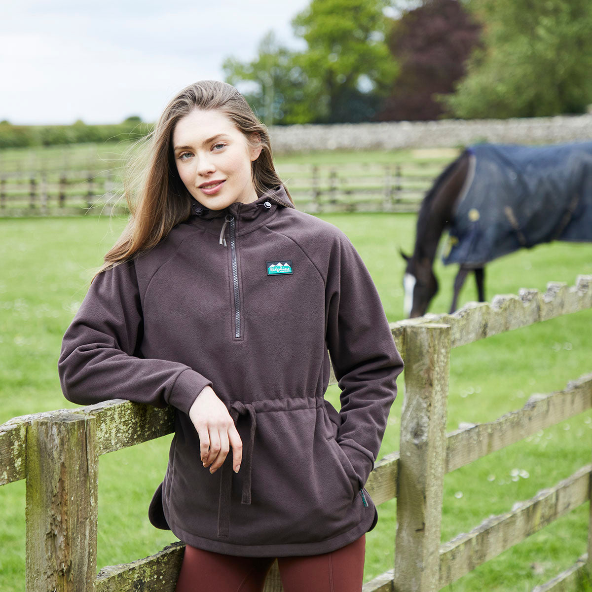 A woman wearing a dark brown half-zip fleece pullover with a drawstring waist and a small teal logo on the chest, standing in a grassy field next to a wooden fence with a horse in the background.