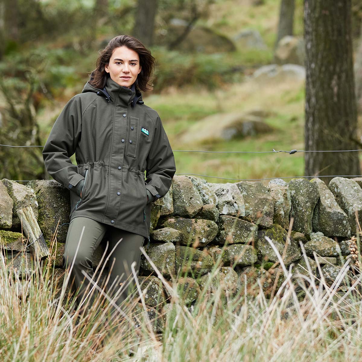 Woman in a green jacket standing next to a stone wall in a natural setting