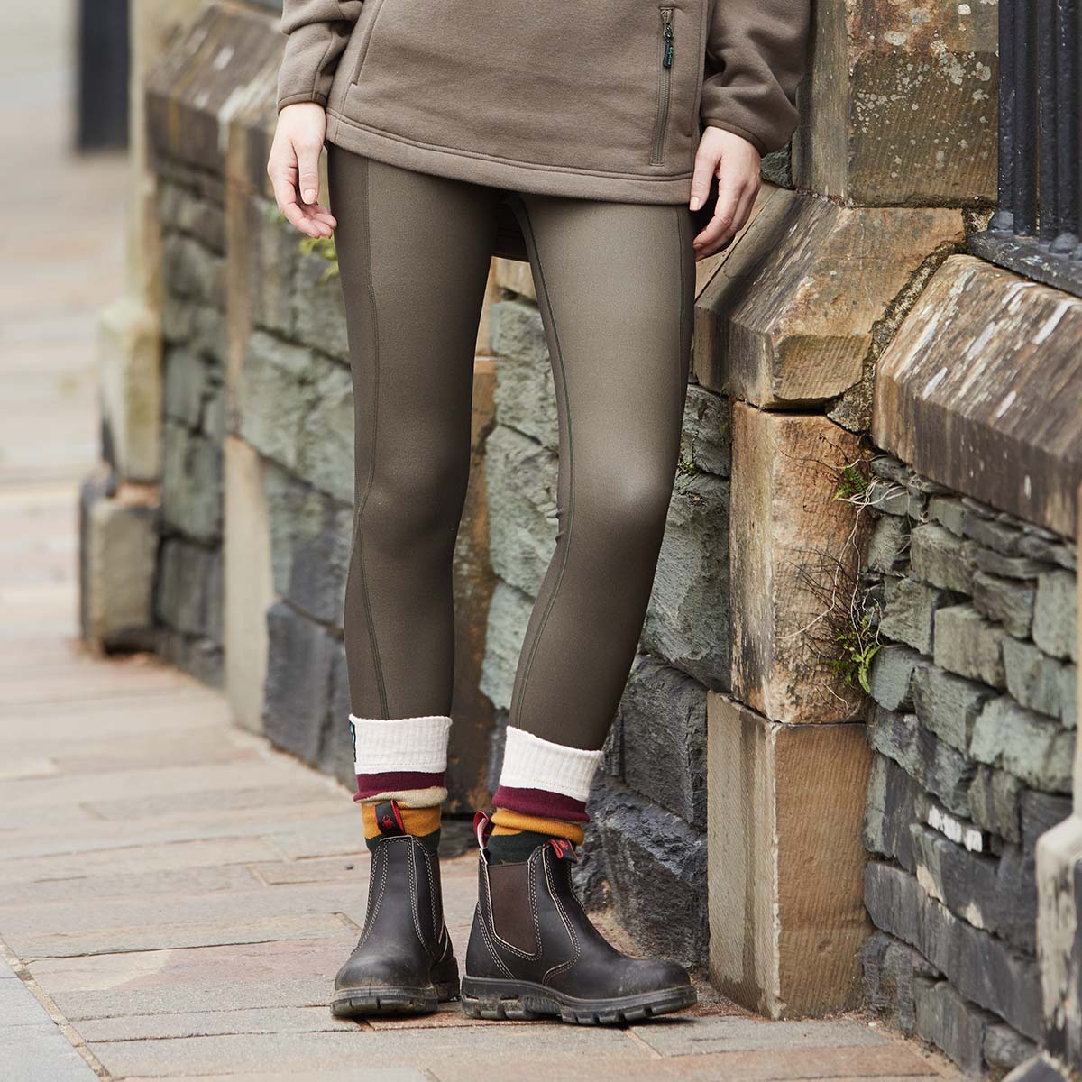Person wearing brown leggings and boots standing against a stone wall.