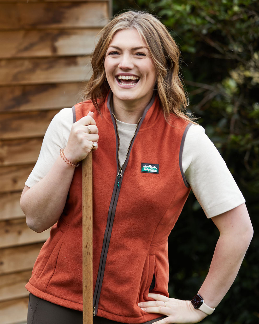 Woman wearing a red vest with a logo, holding a tool outdoors.