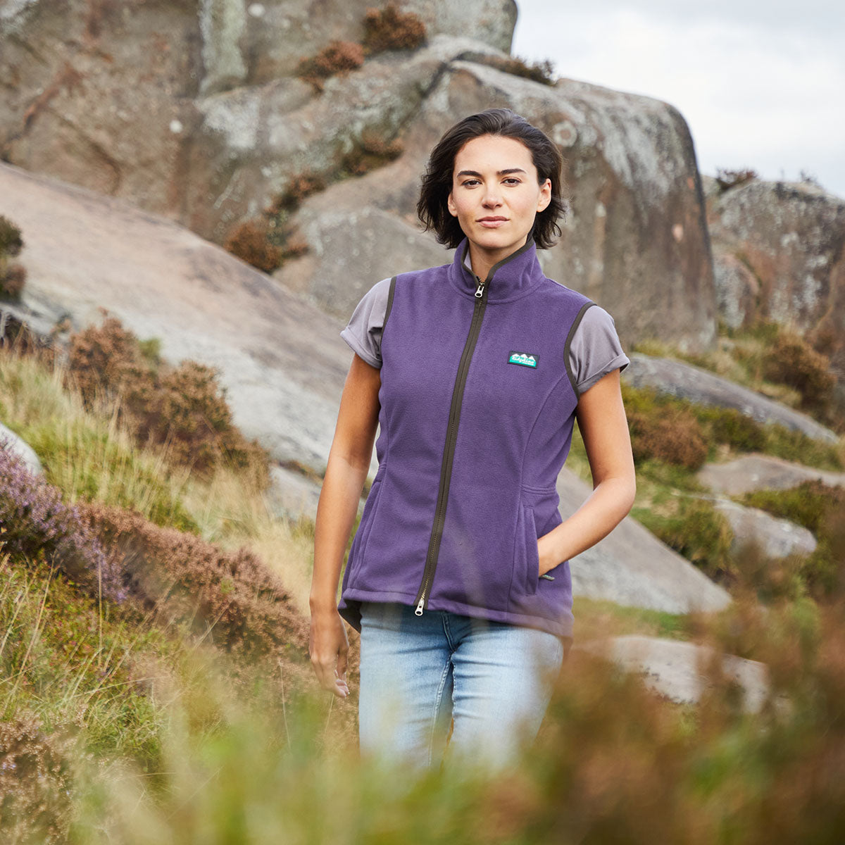 Woman wearing a purple vest with a logo, standing in a natural landscape with rocks and grass.