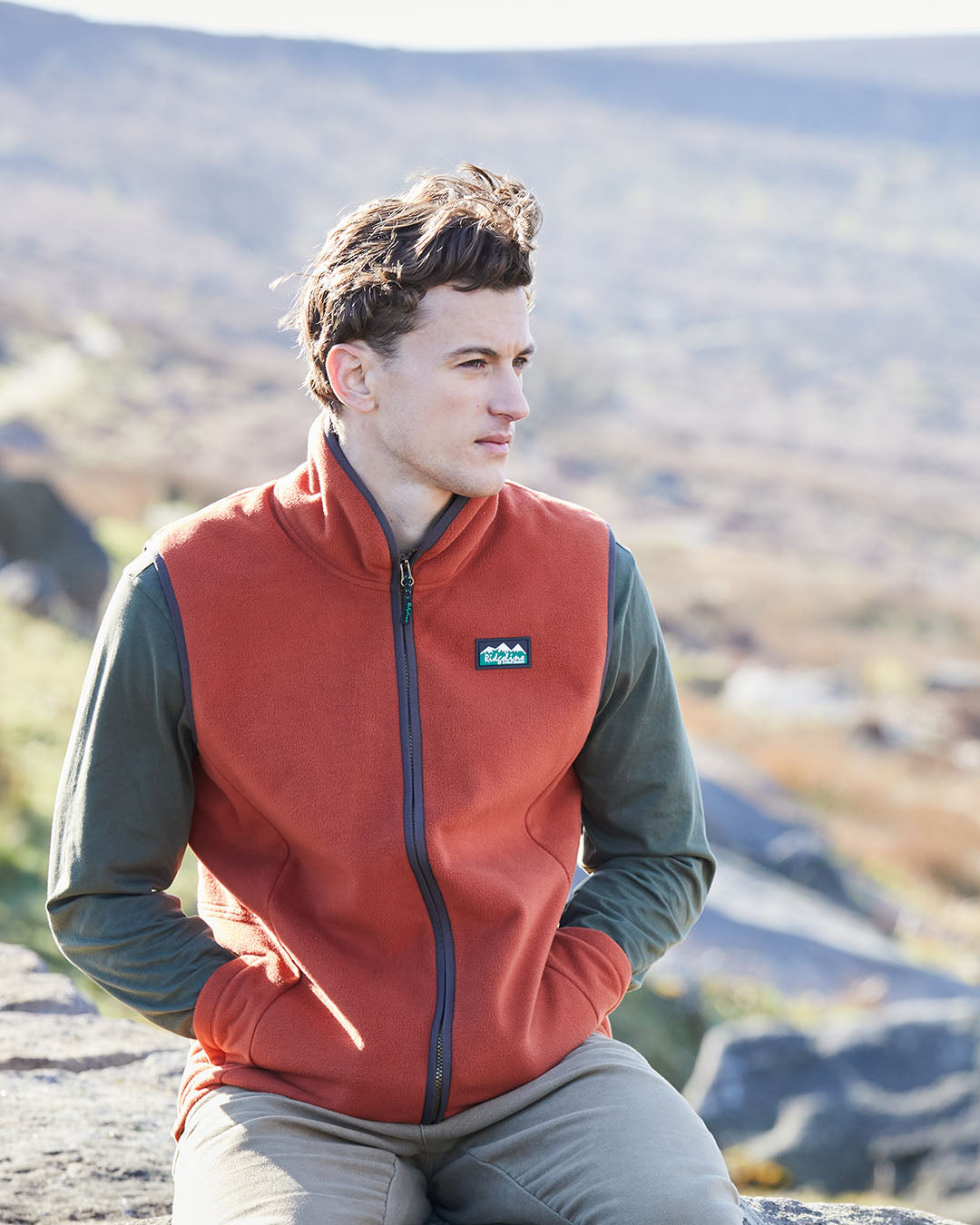 Man wearing a red fleece vest with a visible brand logo, sitting outdoors with mountains in the background.