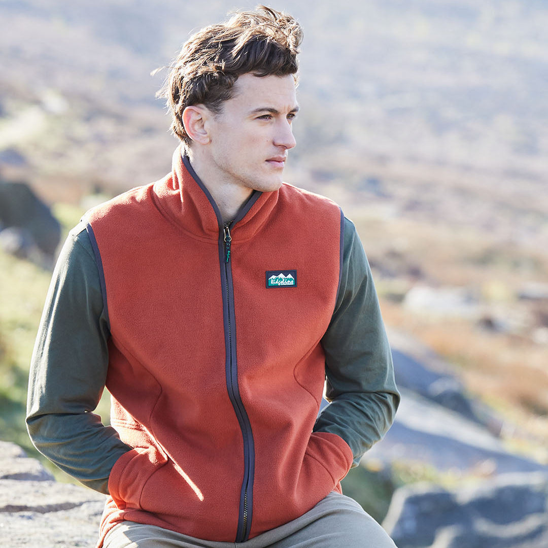 Man wearing a red fleece vest with a visible brand logo, sitting outdoors with mountains in the background.