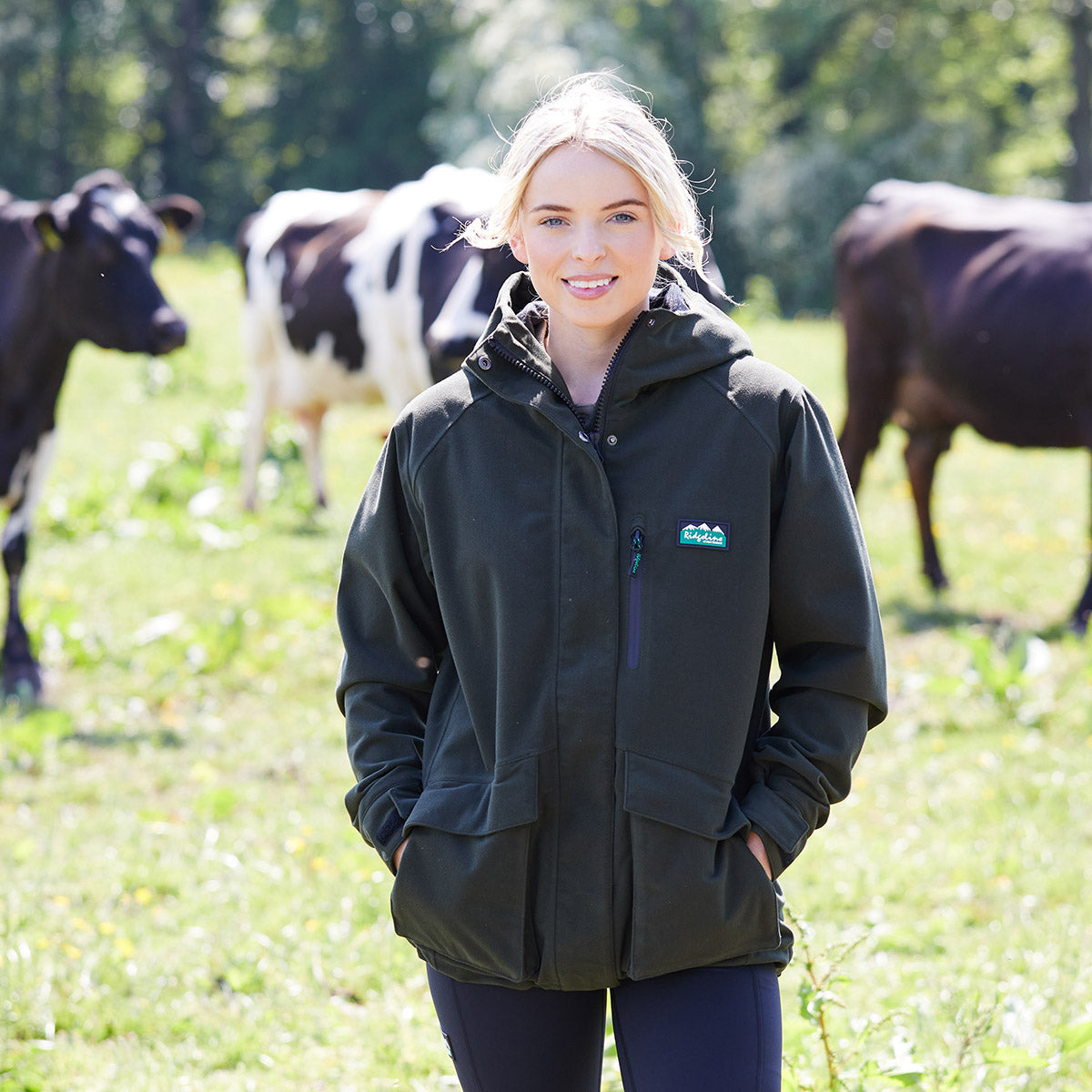 Woman standing in a field with cows wearing a green Ridgeline Clothing branded jacket.