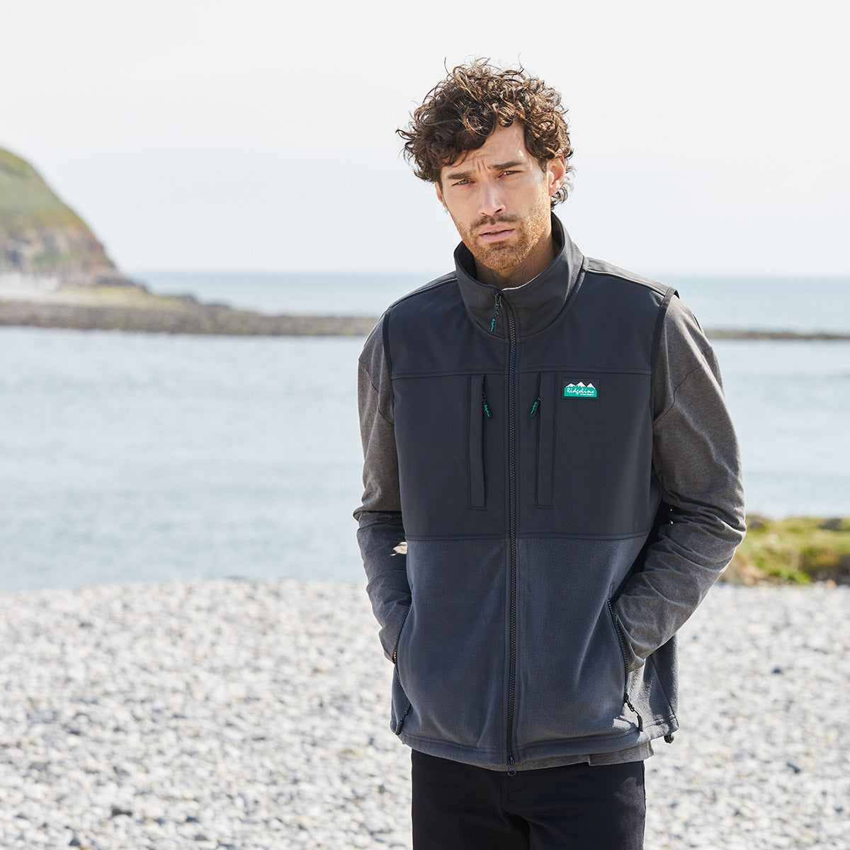 Man wearing a black windproof fleece Gilet with a Ridgeline Clothing logo standing on a pebbly beach.