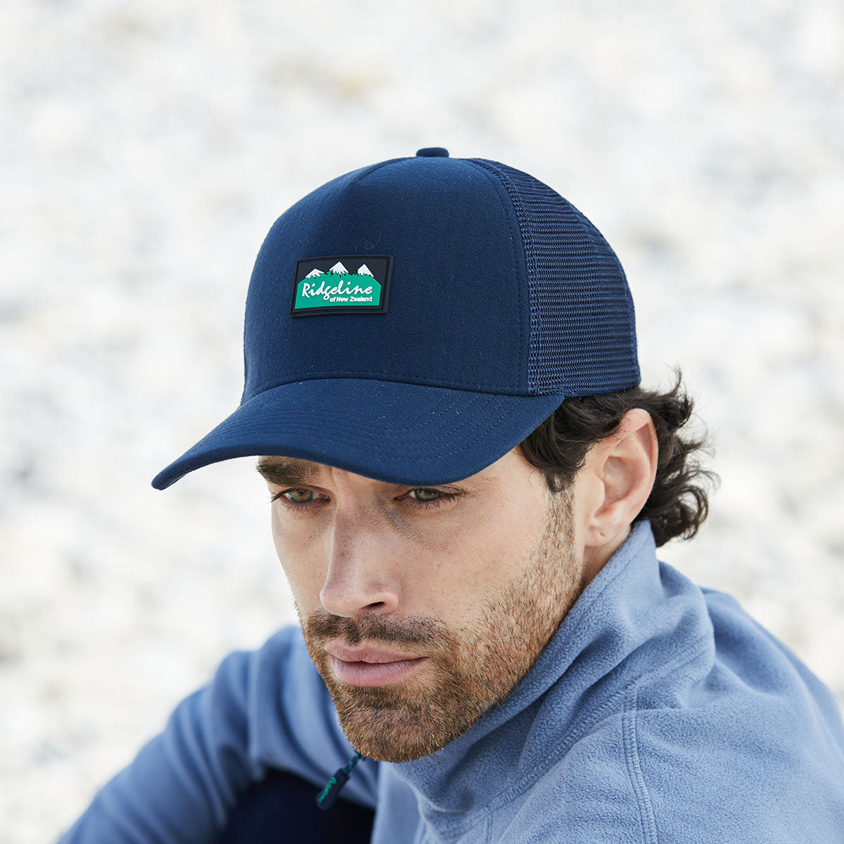 Man wearing a blue cap with a logo, looking downwards against a blurred natural background